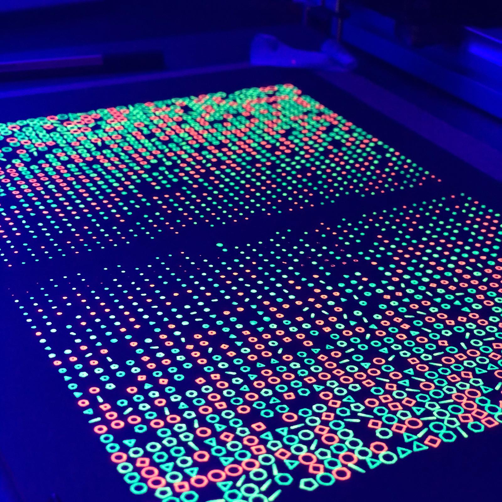 Black paper with a grid of many neon orange, yellow, and green printed shapes, glowing under a dark-purple illumination of a UV light